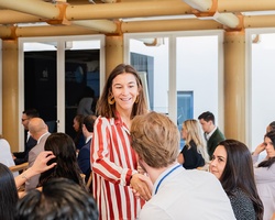 A woman in a striped shirt smiles while greeting a colleague in a busy office setting.