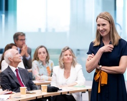 A woman stands smiling at a podium in a seminar room while a group of attentive adults are seated listening to her.