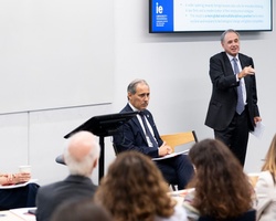 A man speaks at a podium in a lecture hall with attentive audience and two other participants seated.