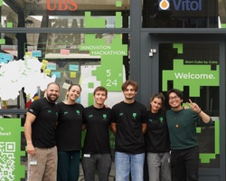 A group of six young adults standing in front of a glass building with green digital-themed decorations.