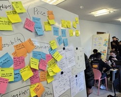 A collaborative workspace featuring a board filled with colorful sticky notes and people brainstorming in the background.