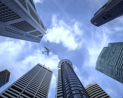 A plane flies above skyscrapers under a partly cloudy sky.