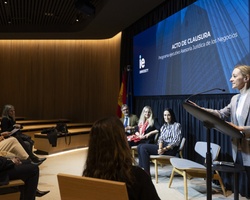 A woman is giving a speech at a podium in front of an audience in a university auditorium.
