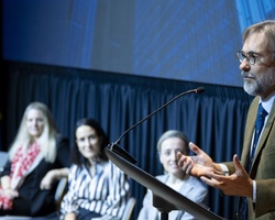 A man is speaking at a podium with four women seated in the background at an event.