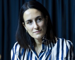 A woman in a striped shirt sits against a dark curtained background, looking thoughtful.