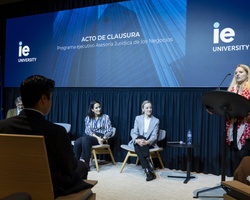 A woman speaks at a podium during a closing ceremony at IE University, with three people seated behind her on stage.