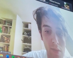 A young man appears on a video call on a computer screen, with a bookshelf in the background.