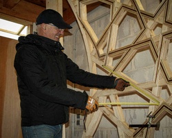 A man is measuring wooden geometric shapes with a tape measure in a workshop.