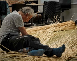 An elderly man sits on the floor working with natural materials.