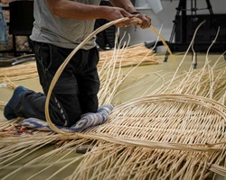 A person is kneeling and weaving with thin, flexible materials on the floor.