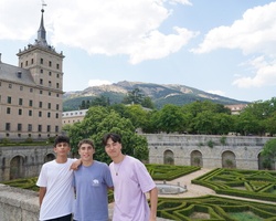 Three young men standing in front of a historic building with gardens and mountains in the background.