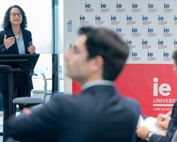 A speaker presents at a law school event with attentive audience members in the background.