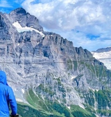 A person in a blue jacket standing in front of majestic mountains under a blue sky.