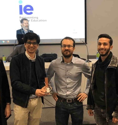 A group of five people, one holding an award, posing in a seminar room with a presentation on 'Reinventing Education' in the background.