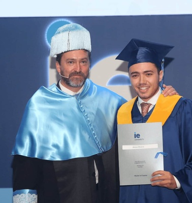 A young graduate in blue and yellow gown holding a diploma stands next to an academic in blue ceremonial robes during a graduation ceremony.