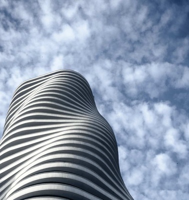 A modern white cylindrical building with horizontal ribbed patterns, set against a bright blue sky with scattered clouds.