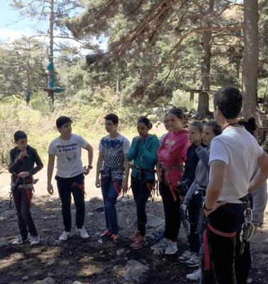 A group of young people equipped with climbing gear standing in a forest, listening to an instructor.