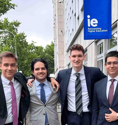 Four young men in suits posing together in front of a building with a sign that reads 'IE Reinventing Higher Education'.