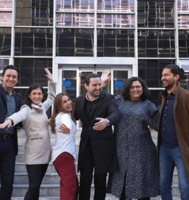 A group of six people joyfully posing together in front of a modern building, some waving.