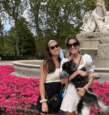 Two women smiling with a dog in front of a flower bed and a statue in a park.