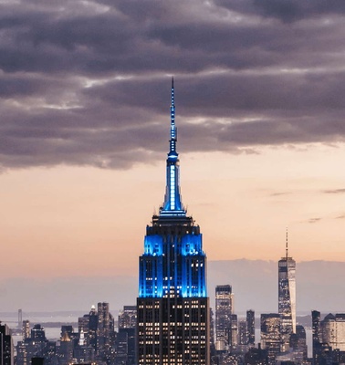 A view of the Empire State Building illuminated in blue at dusk with a cityscape background.