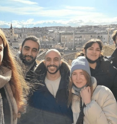 A group of six friends taking a selfie with a cityscape in the background during daytime.