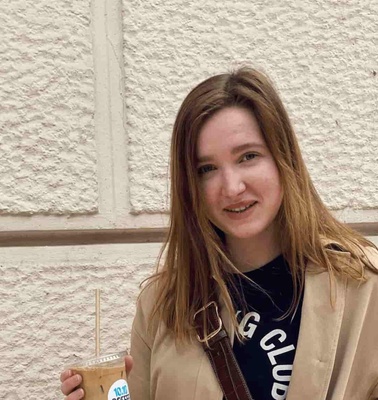 A young woman smiles while holding a cold beverage, standing in front of a textured wall with a sign.