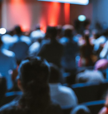 A blurred image of an audience at a conference with a speaker in the background.
