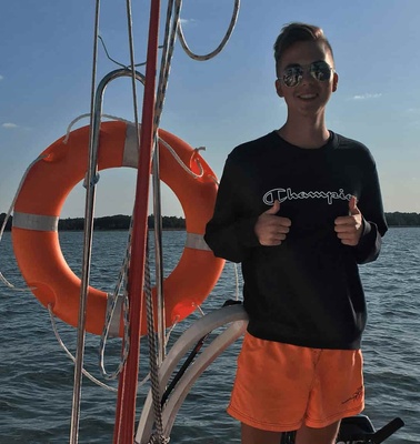 A young man stands on a boat holding a mug, with a lifebuoy and lake in the background.