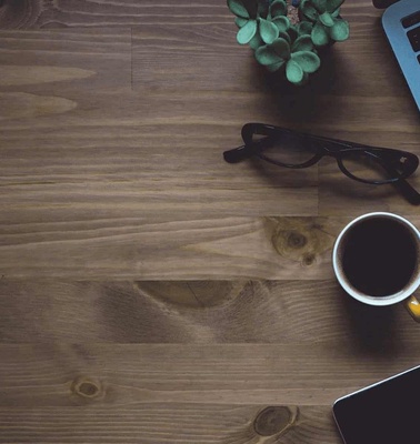 A workspace with a laptop, coffee cup, glasses, and a small potted plant on a wooden table.