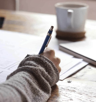 A person writing on a paper at a wooden table with a cup of coffee beside them.