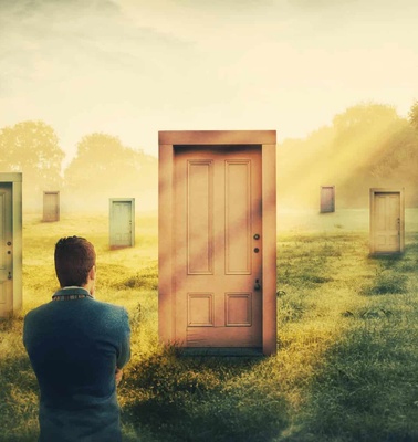 A man stands in a grassy field looking at multiple doors under a sunlit sky.