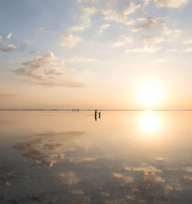 A serene sunset over a vast reflective water surface with a few people walking in the distance.