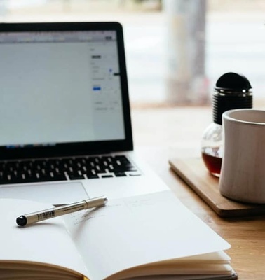 A cozy workspace with an open laptop, a notebook, a pen, and a cup of coffee on a wooden table.