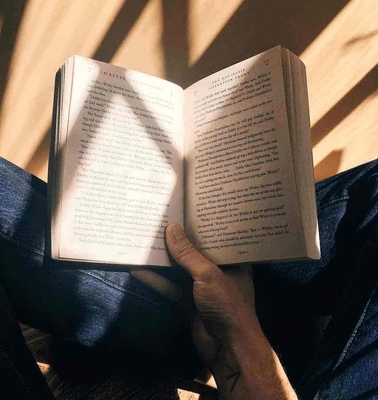 A person sitting with crossed legs holding an open book in a sunlit room.