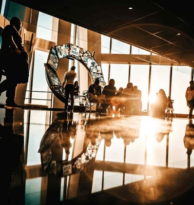 Silhouetted figures in a modern building interior with a prominent glass clock during a sunset, creating reflections on the shiny floor.