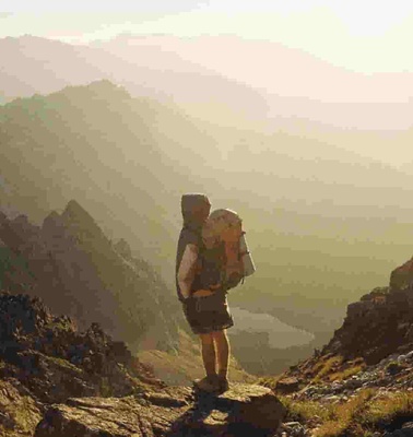 A hiker stands at a mountain pass looking over a layered mountain range during sunset.