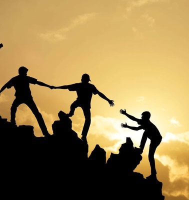 Silhouette of people helping each other to climb up a rocky hill against a sunset background.