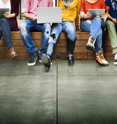A group of young adults sitting on a bench using various digital devices.