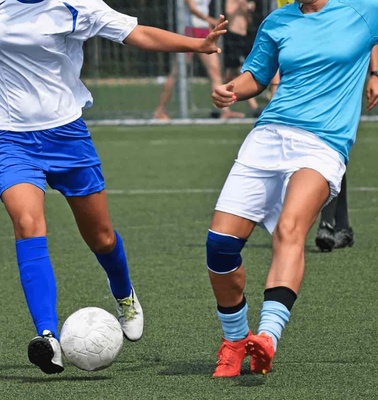 Two female soccer players in a match, one wearing a blue jersey and the other in light blue, competing for the ball on a grass field.