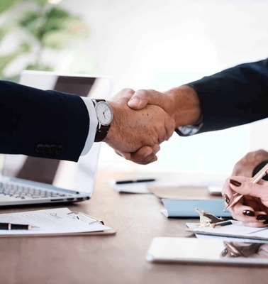 Two professionals shaking hands over a desk with laptops and documents, signifying a business agreement.