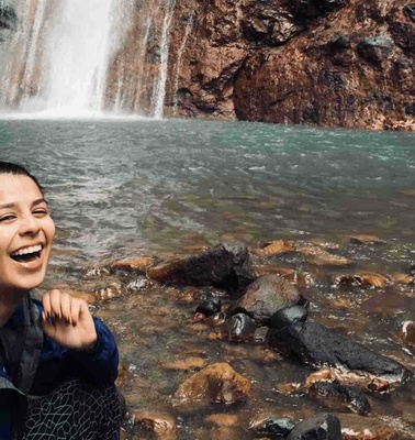 A woman smiling joyfully near a waterfall with clear waters and rocky surroundings.