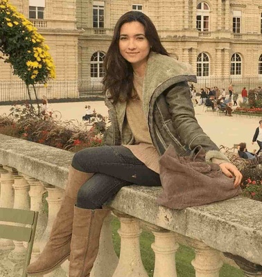 A woman sits on a stone railing in front of a historic building surrounded by flowering plants.