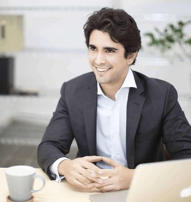 A young man in a suit sits at a desk with a laptop and a coffee cup, smiling in an office setting.