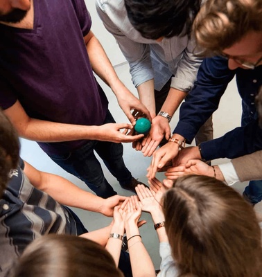 A group of people stands in a circle, placing their hands together with one person holding a small green object.