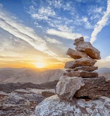 A stack of stones balanced on a rocky mountain summit during sunset with a cloudy sky in the background.