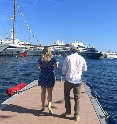 A man and a woman walking on a dock overlooking luxury yachts in a sunny marina.