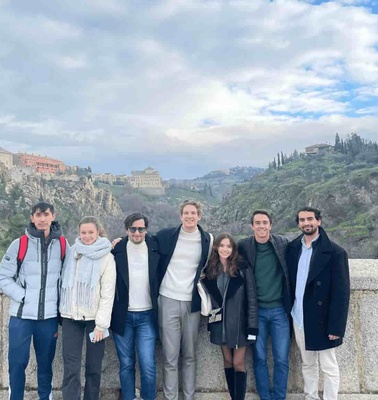 A group of seven people posing together on a bridge with a historic cityscape in the background.