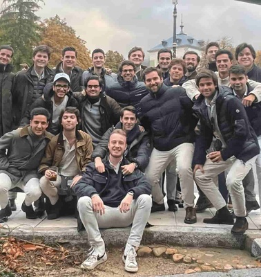 A group of young men posing together outdoors in a public place, smiling and embracing each other, with trees and a cityscape in the background.