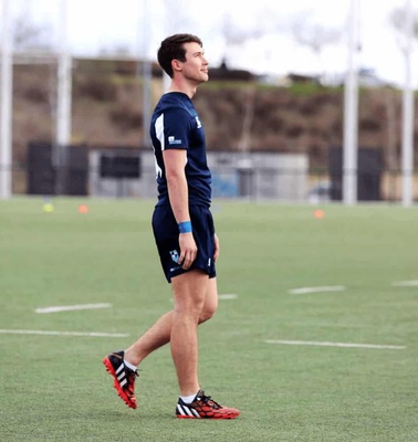 A young male athlete in a blue uniform standing on a soccer field, looking to the side, with other players training in the background.